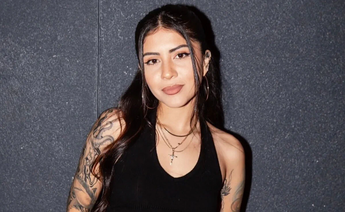 Nina J with long dark hair and tattooed arms in front of a grey wall in a promo portrait