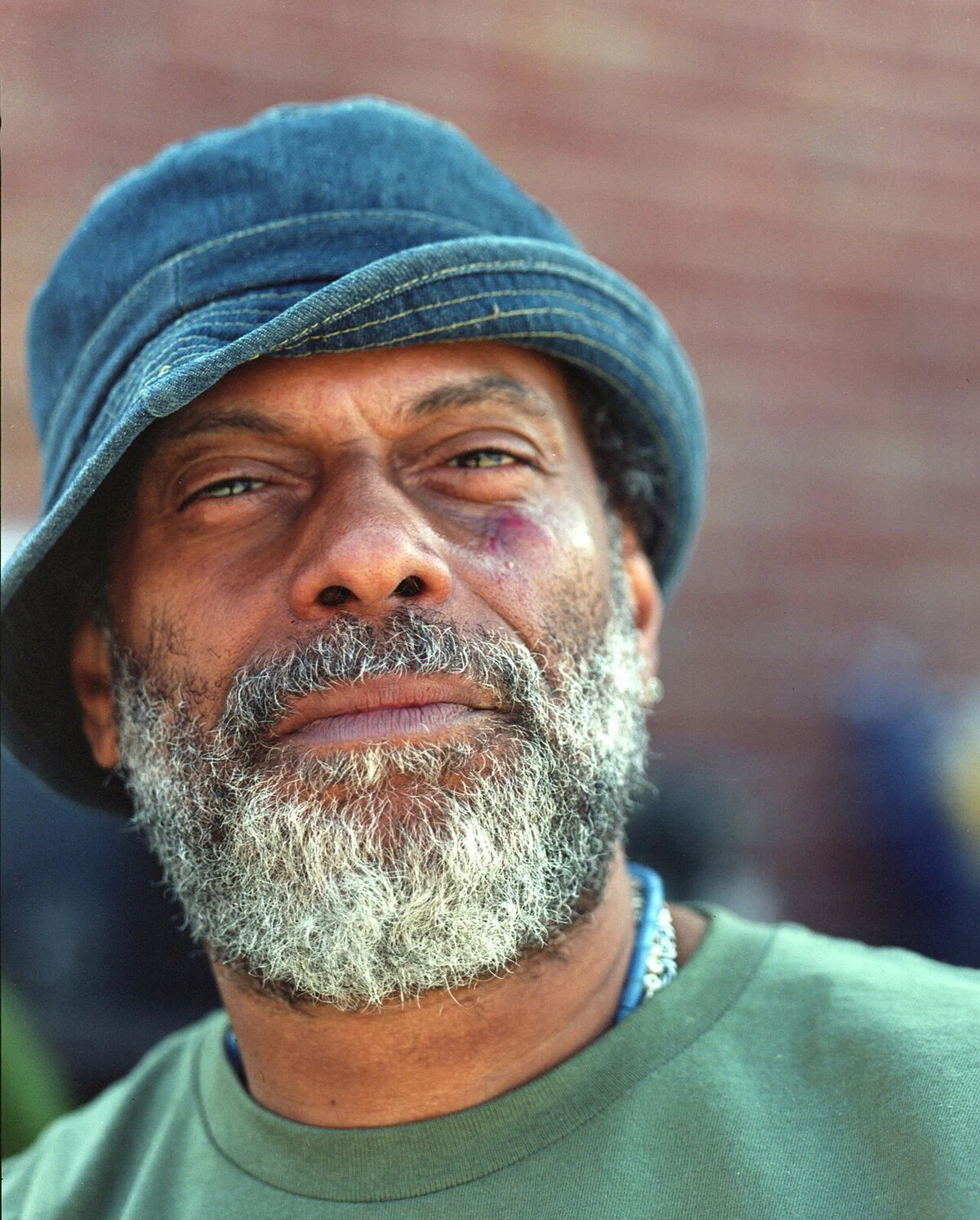 Portrait of Theo Parrish, a bearded man in a denim bucket hat and green shirt in front of a brick wall