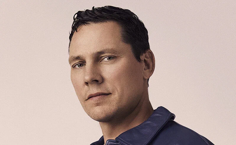 Tiesto in a head-and-shoulders promo portrait against a pale pink backdrop