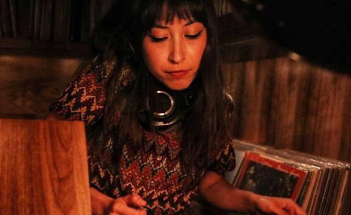 Xica Soul at the decks with headphones around her neck, looking down at a crate of vinyl records