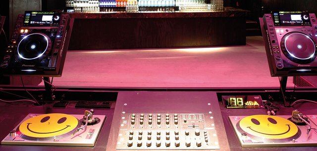 A DJ booth with two CDJ players and a mixer, each deck topped with a smiley-face mat