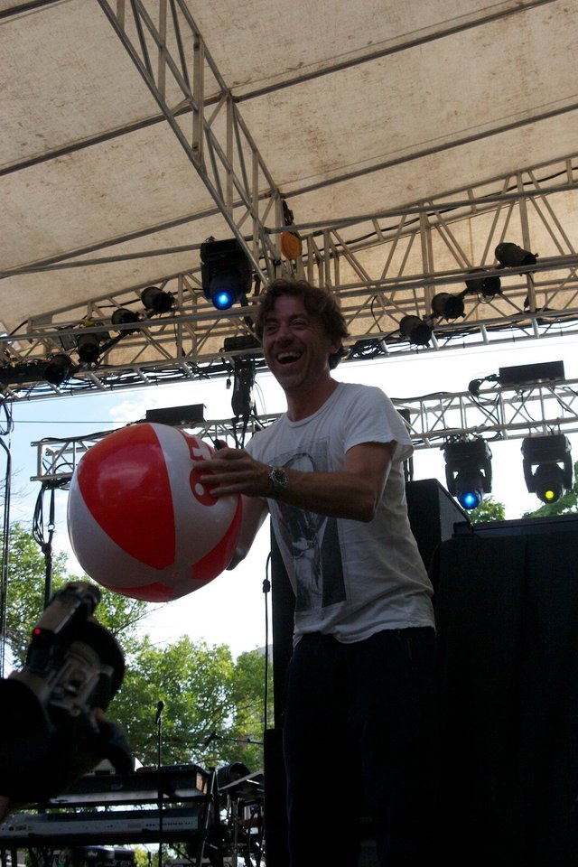 Benny Benassi laughing on an outdoor stage under a lighting truss while holding a red and white beach ball