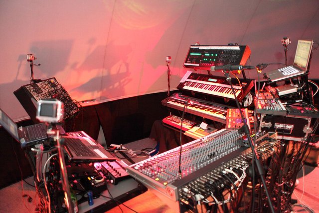 A stage rig of synthesizers, keyboards and electronic gear lit in red and pink light, with no one at the controls