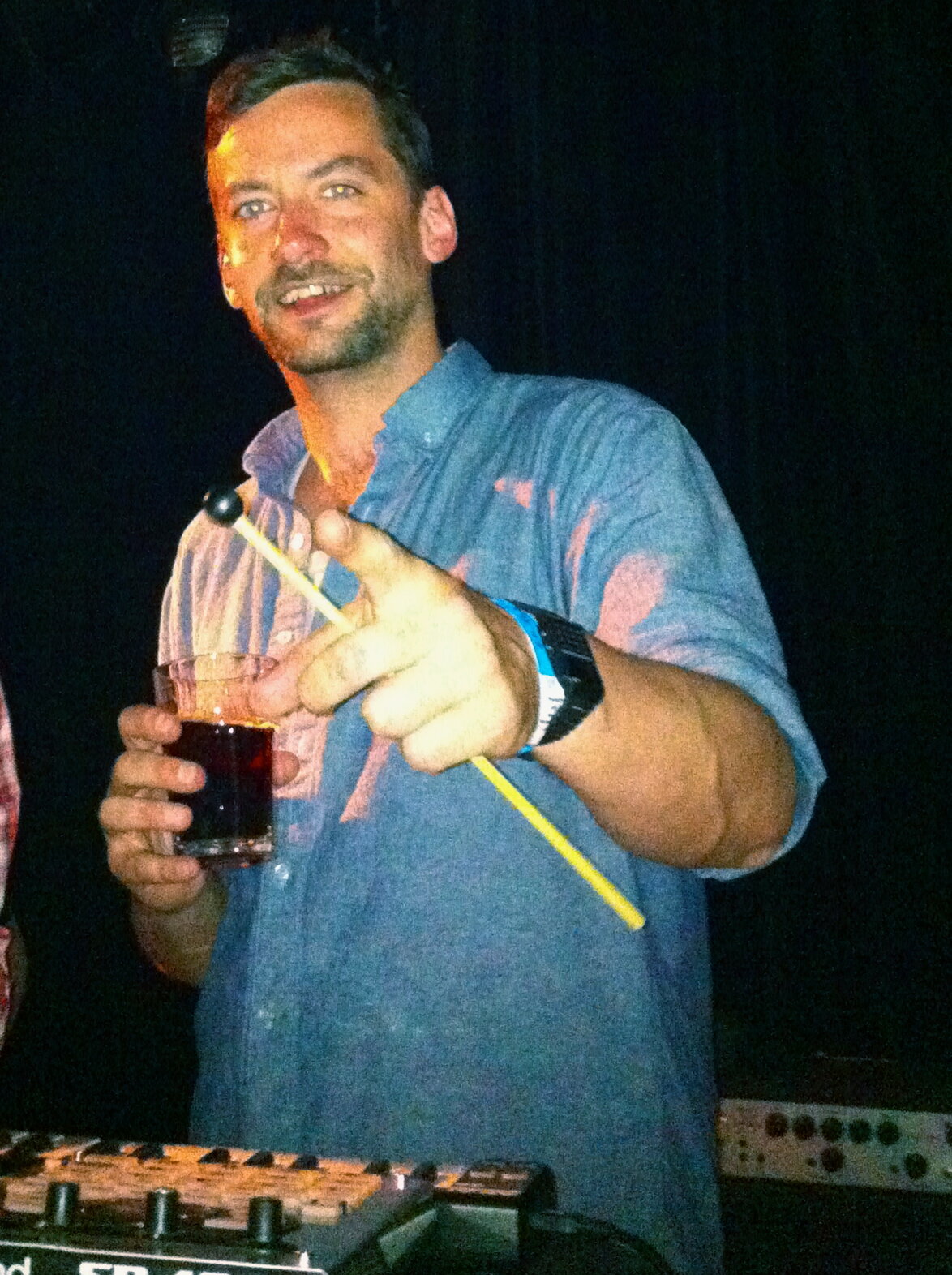Bonobo smiling at the camera behind his live gear, holding a drink and a mallet