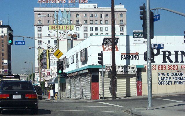 Corner of 7th Street and Bixel Street in Los Angeles with traffic signals, street signs and a mid-rise apartment building behind a row of storefronts