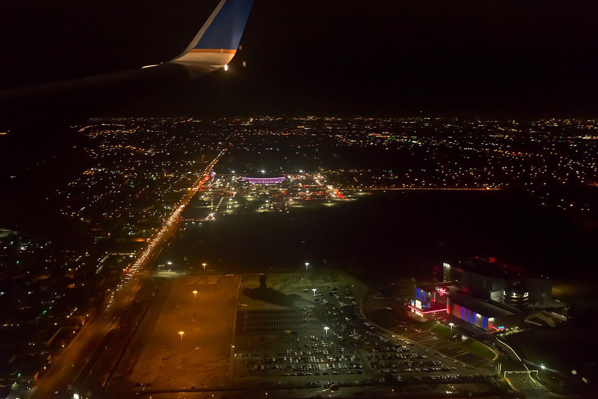 Night aerial view from a plane window of Inglewood's lit streets with the Kia Forum glowing purple in the distance