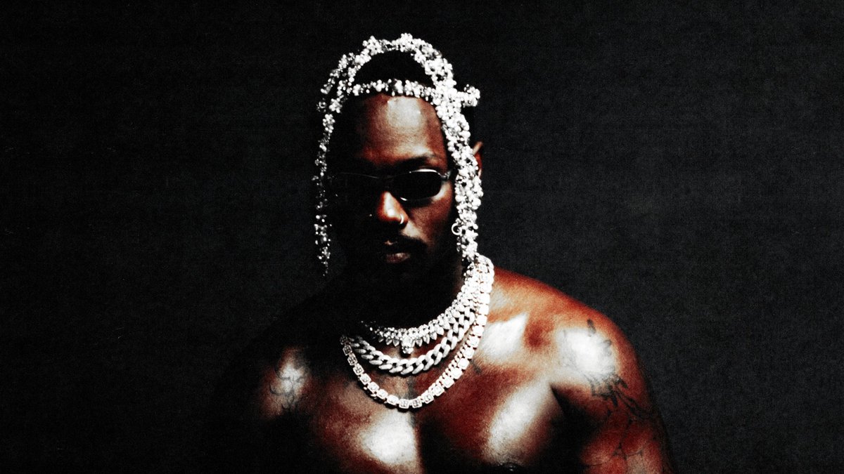 Channel Tres, shirtless in dark sunglasses, wearing a jeweled headpiece and stacked silver chains against a black background