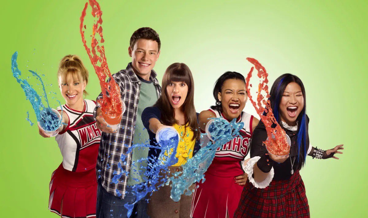 Five smiling young people, two in red and white cheerleader uniforms, holding up cups that spray blue and orange liquid against a green background