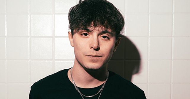 Portrait of bass producer Costa in a black t-shirt and chain against a white tiled wall