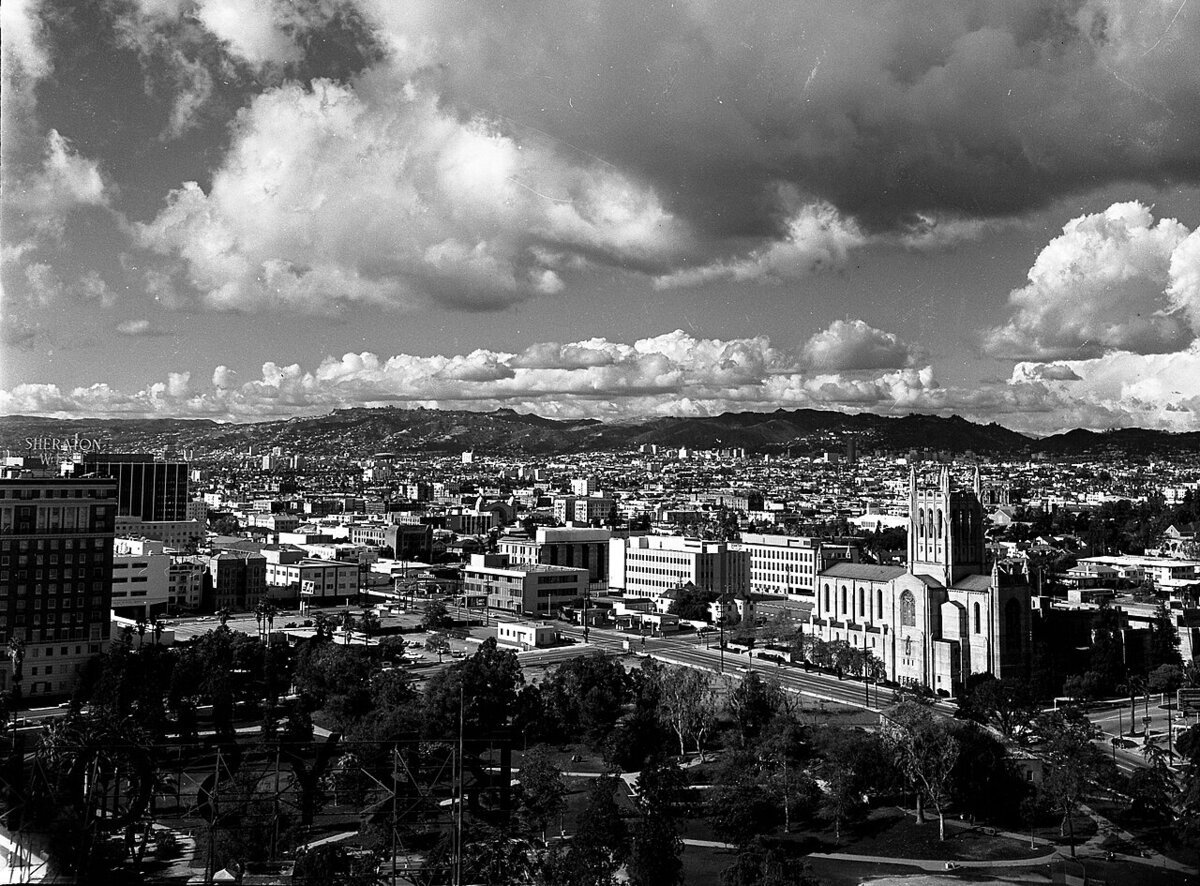 Archival black-and-white view over Los Angeles rooftops toward the hills, with a stone church tower and park in the foreground