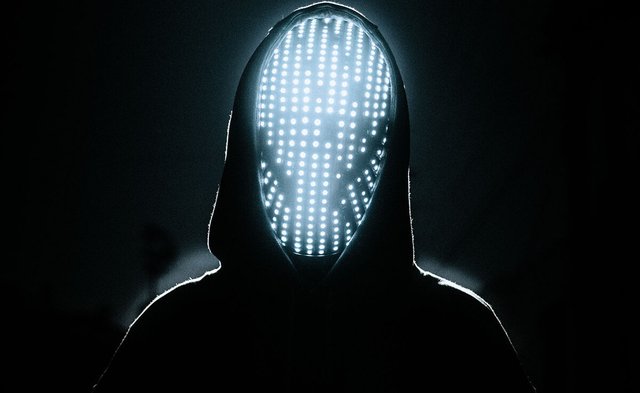 Hooded figure facing the camera with a face covered by a glowing grid of white LED lights against a black background