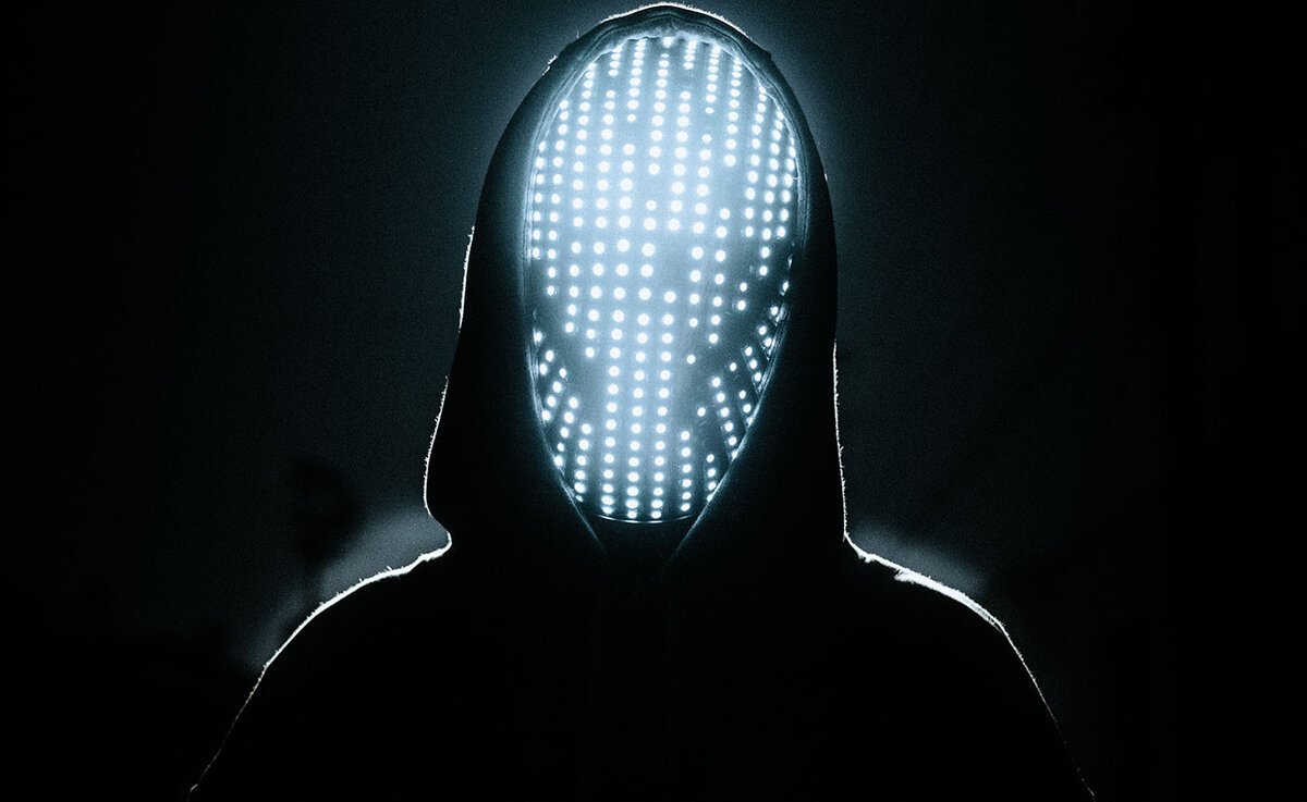 Hooded figure facing the camera with a face covered by a glowing grid of white LED lights against a black background