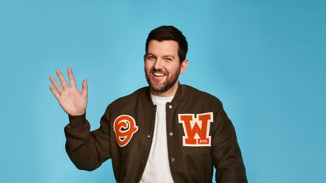 Dillon Francis smiling and waving in a brown varsity jacket against a bright blue backdrop