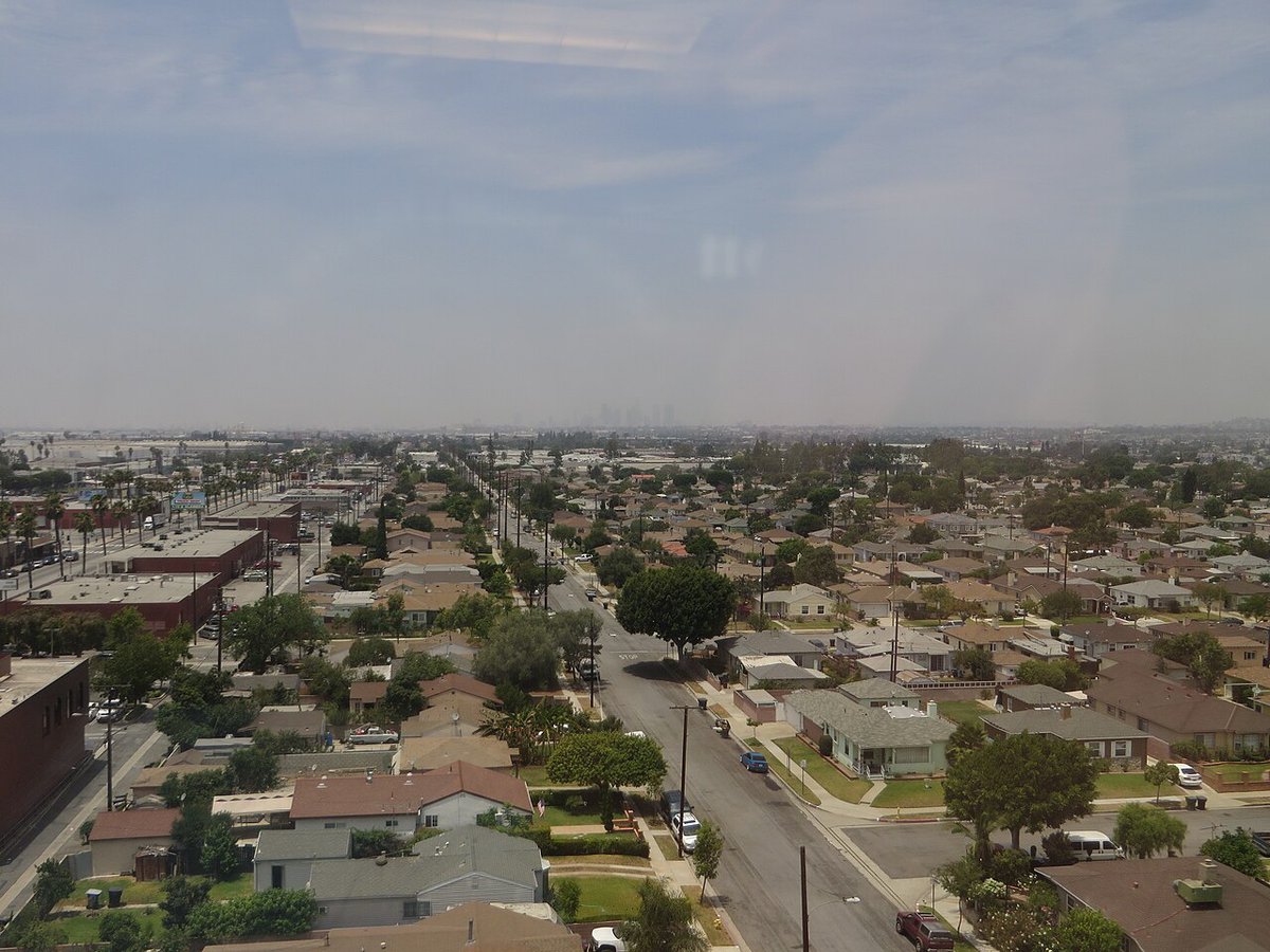 Aerial view over the low rooftops and palm-lined streets of Commerce, with the downtown Los Angeles skyline faint on the hazy horizon