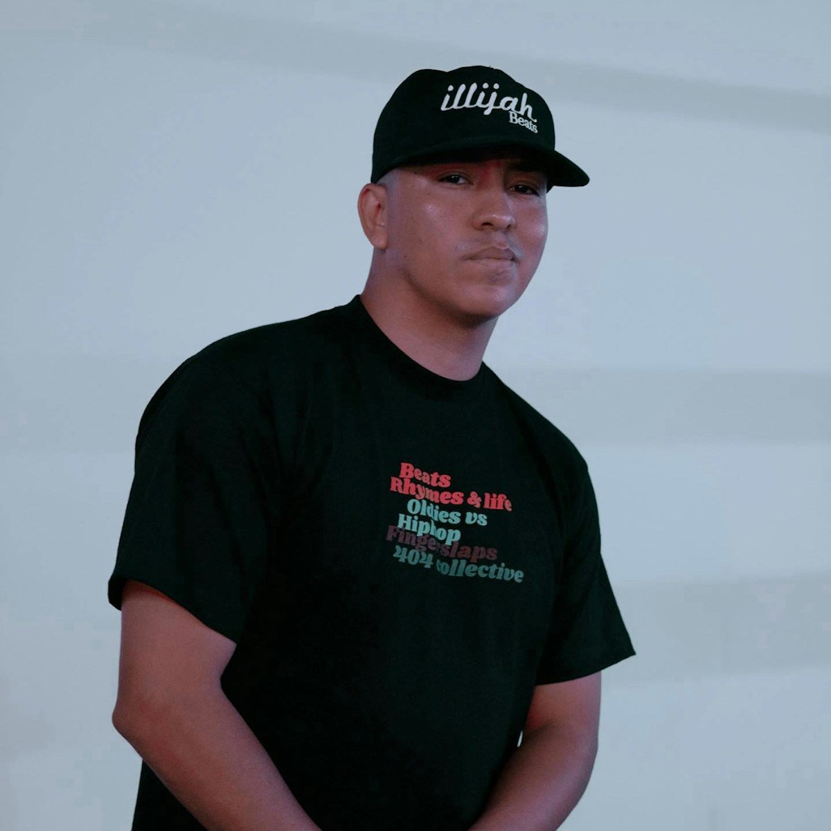 DJ Illijah Beats in a black cap and black t-shirt standing against a pale wall