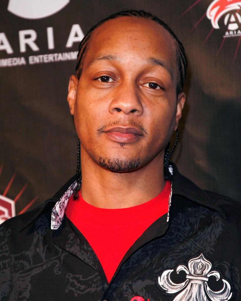 Close-up portrait of DJ Quik in a black patterned shirt over a red tee, standing in front of a dark step-and-repeat backdrop