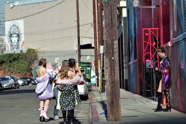 Group in sparkly, fur-trimmed outfits on an Arts District street beside a neon-lit wall and a mural