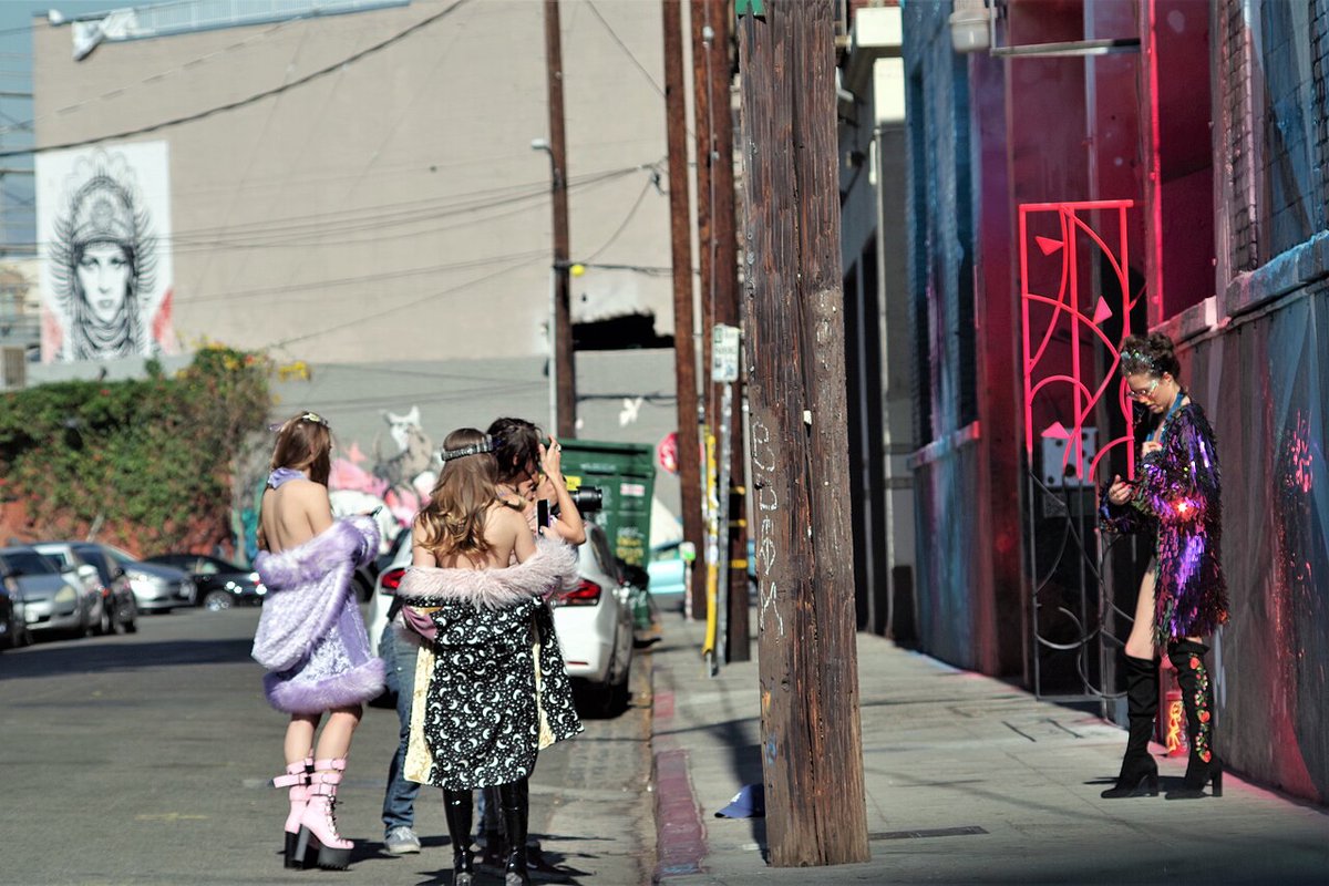 Group in sparkly, fur-trimmed outfits on an Arts District street beside a neon-lit wall and a mural