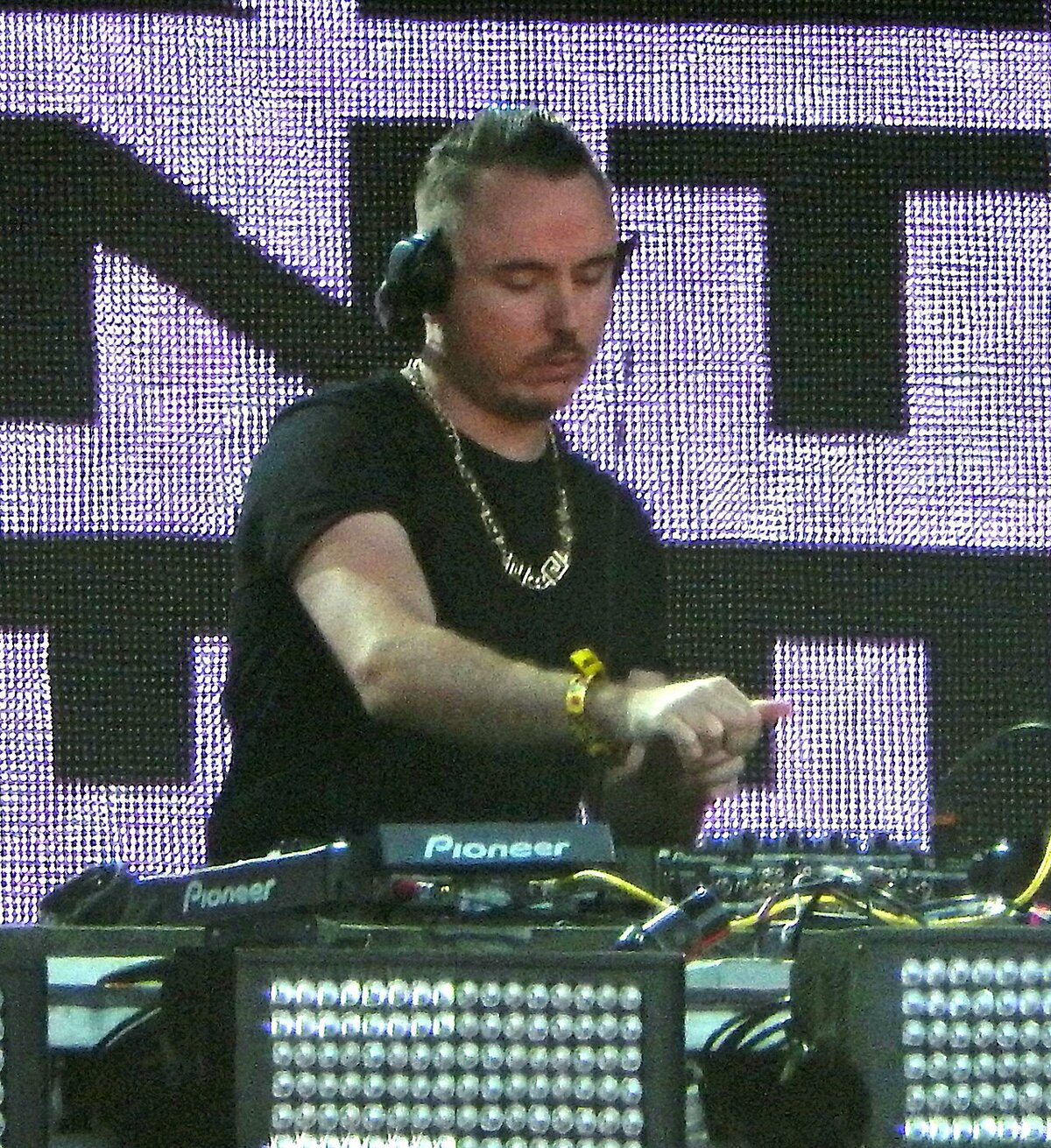 Duke Dumont in headphones and a black shirt behind Pioneer DJ gear in front of a purple LED screen