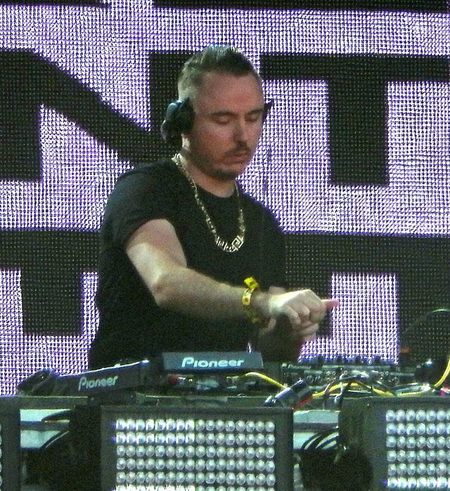 Duke Dumont in headphones and a black shirt behind Pioneer DJ gear in front of a purple LED screen