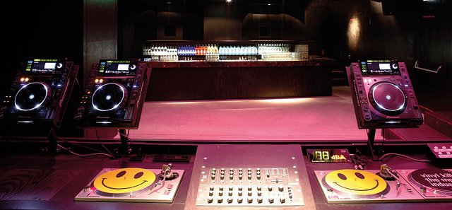 An empty DJ booth with two CDJ players, a mixer and smiley-face slipmats under pink lighting