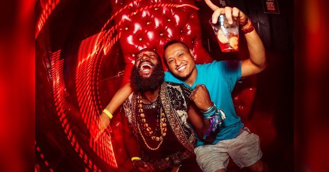 Two smiling clubgoers posing on a red tufted couch under streaks of red light