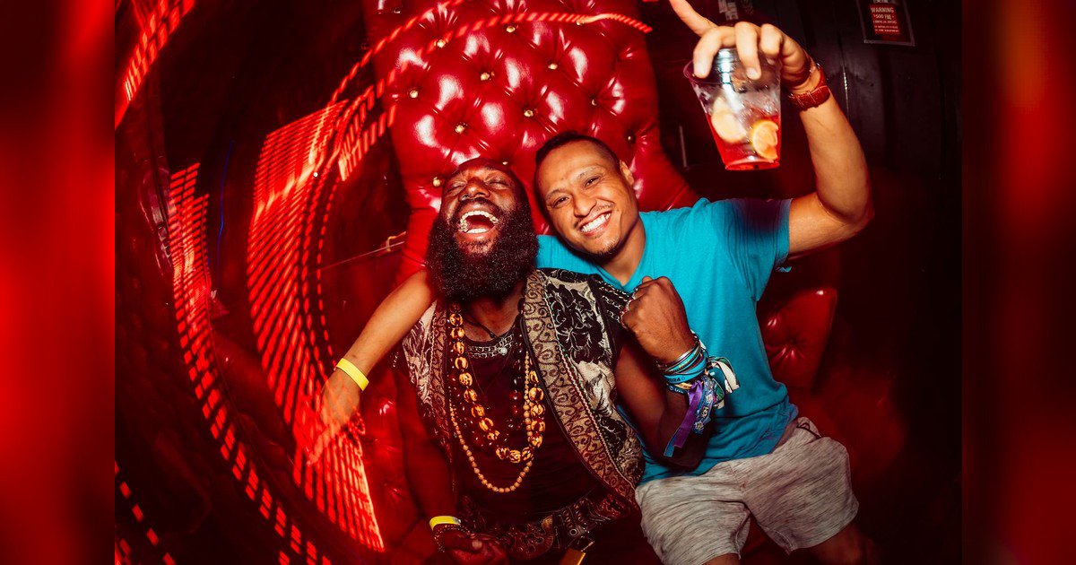 Two smiling clubgoers posing on a red tufted couch under streaks of red light