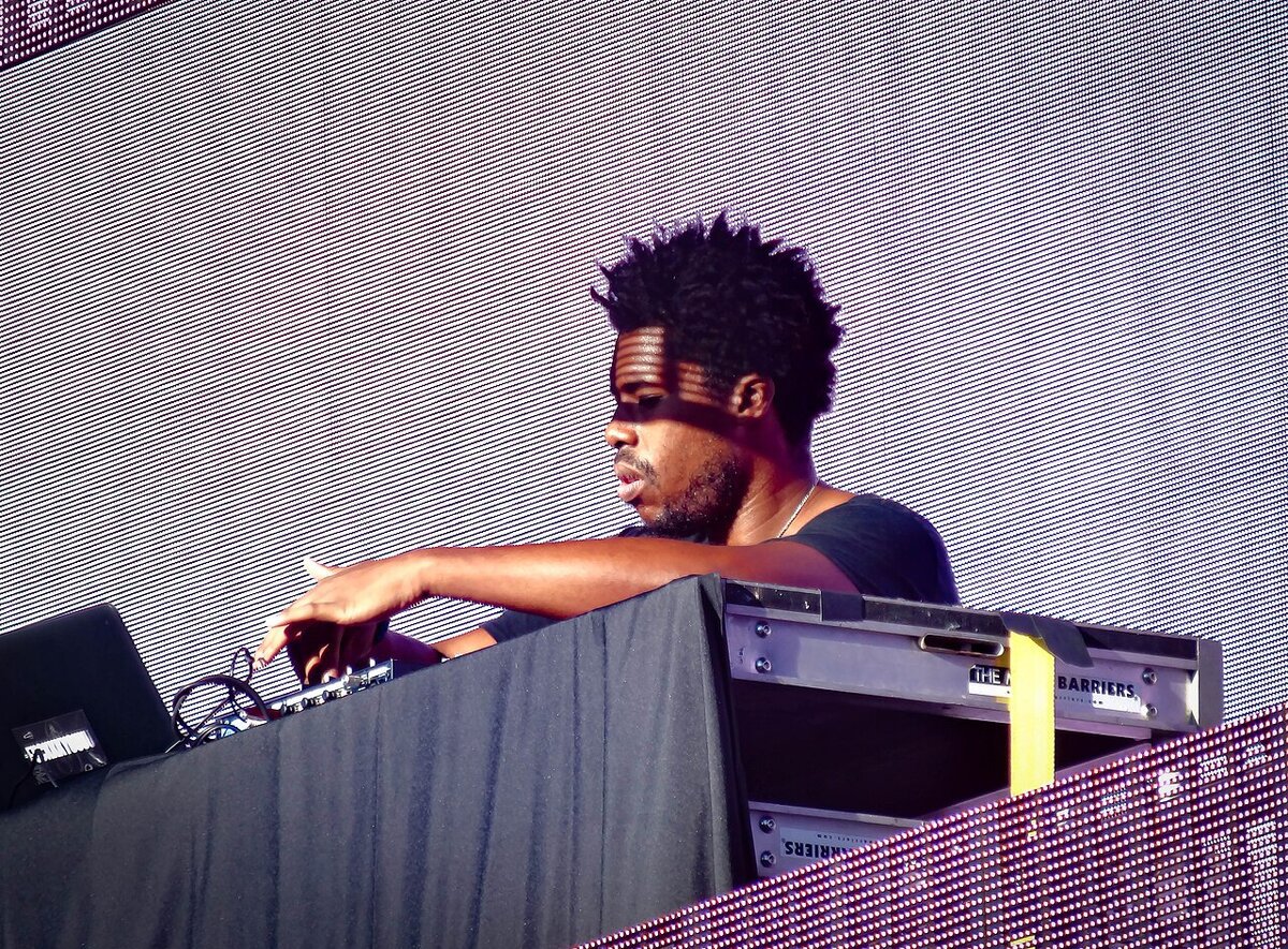 Flying Lotus performing behind a DJ booth in front of a large LED video wall at an outdoor festival