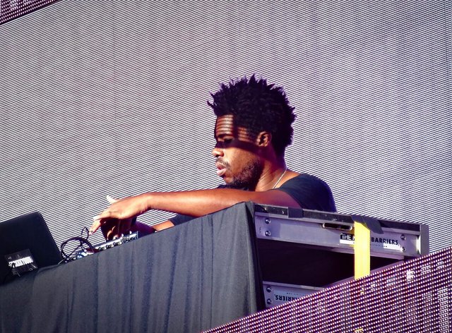 Flying Lotus performing behind a DJ booth in front of a large LED video wall at an outdoor festival