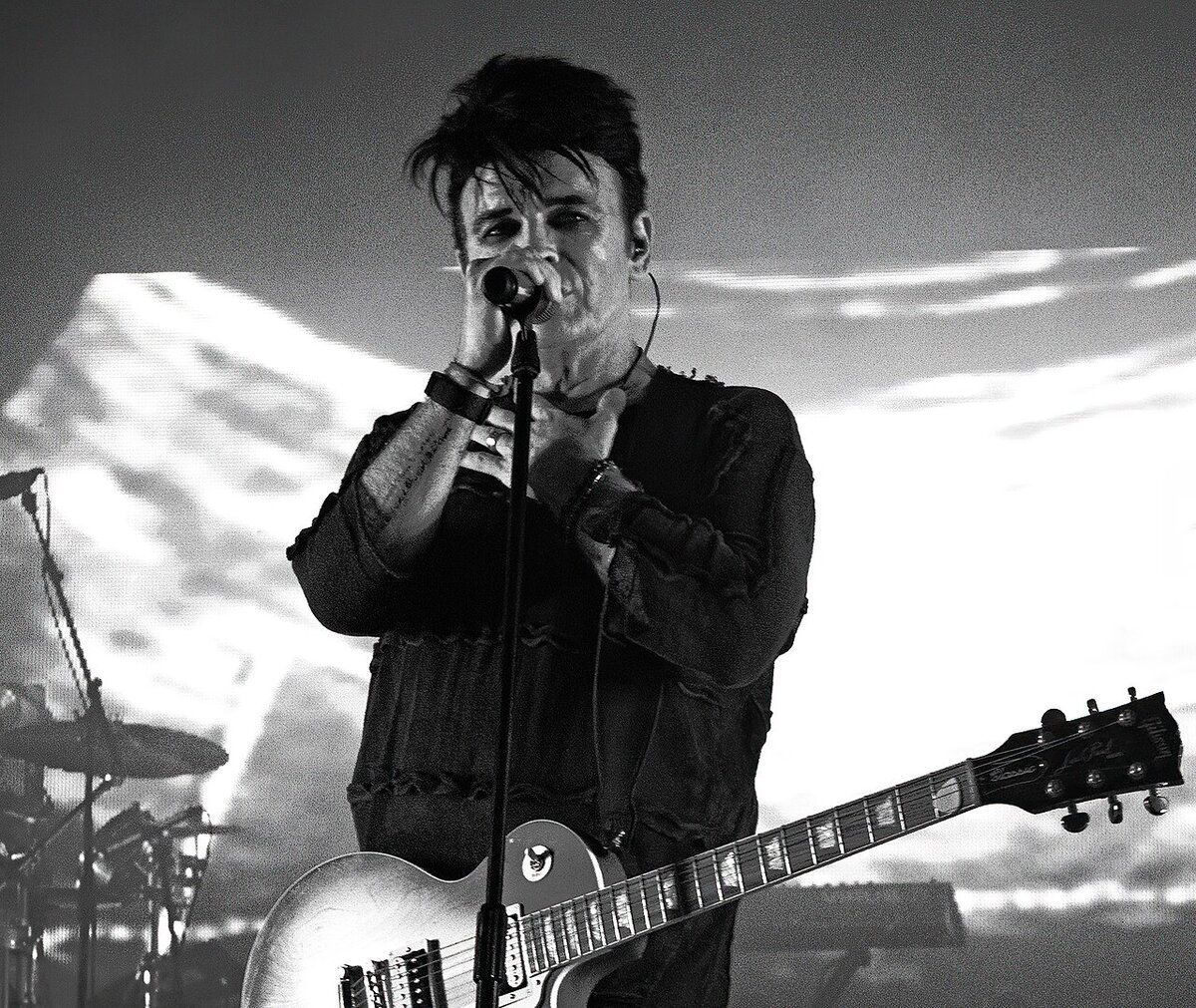 Black and white photo of Gary Numan singing into a microphone, a guitar slung across his chest, in front of a projected cloudscape