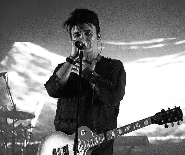 Black and white photo of Gary Numan singing into a microphone, a guitar slung across his chest, in front of a projected cloudscape