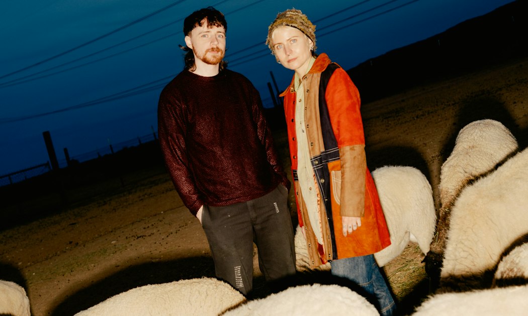 Two people standing at dusk among a flock of sheep, promotional photo for Green-House