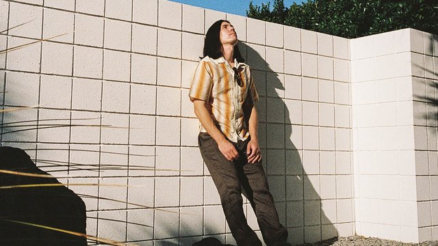 Gryffin, with shoulder-length dark hair and an orange striped shirt, leaning against a white cinder block wall in bright sun