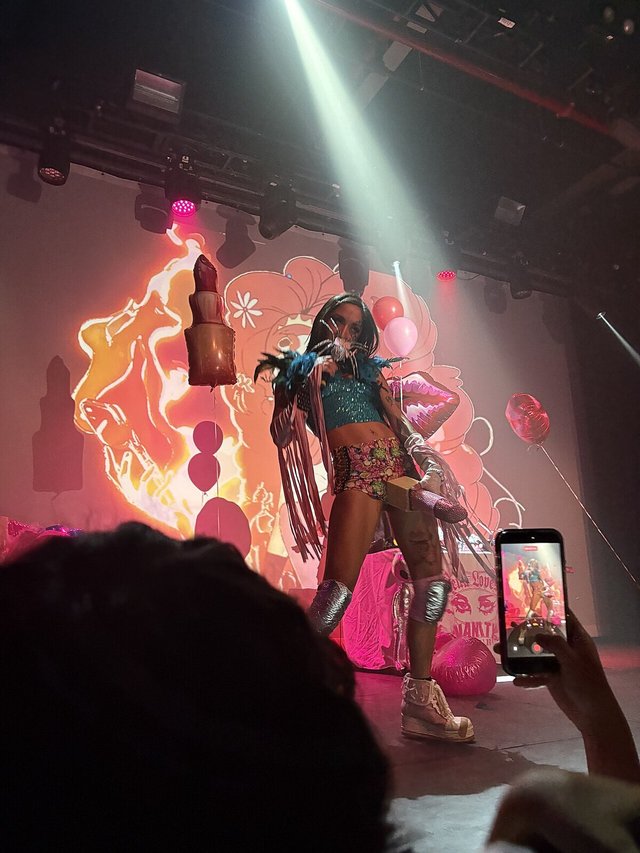 Isabella Lovestory performing on stage in a teal top and fringe jacket, lit by a spotlight with pink balloons and projected artwork behind her