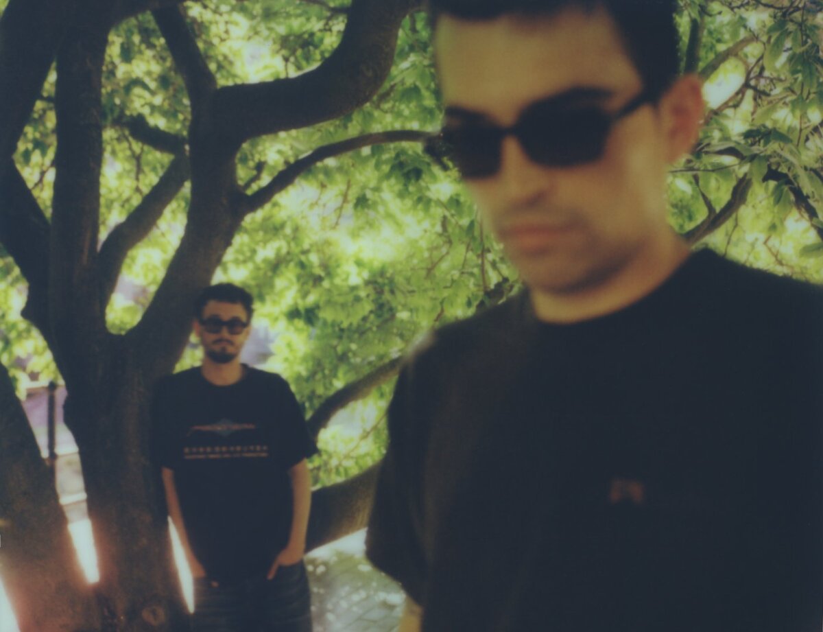Two men in dark sunglasses and black t-shirts outdoors under a leafy tree, one blurred in the foreground and one standing behind