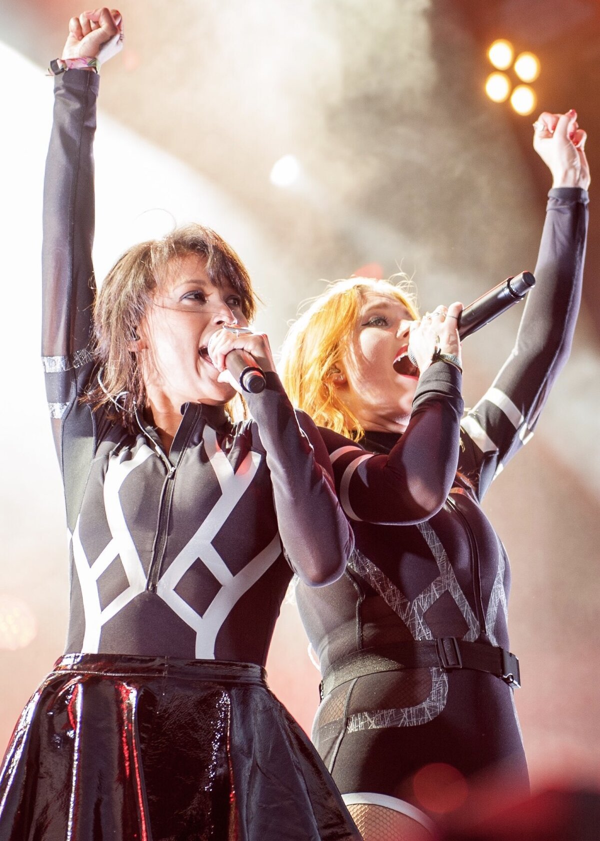 Two vocalists with arms raised, singing into microphones on a hazy, lit festival stage