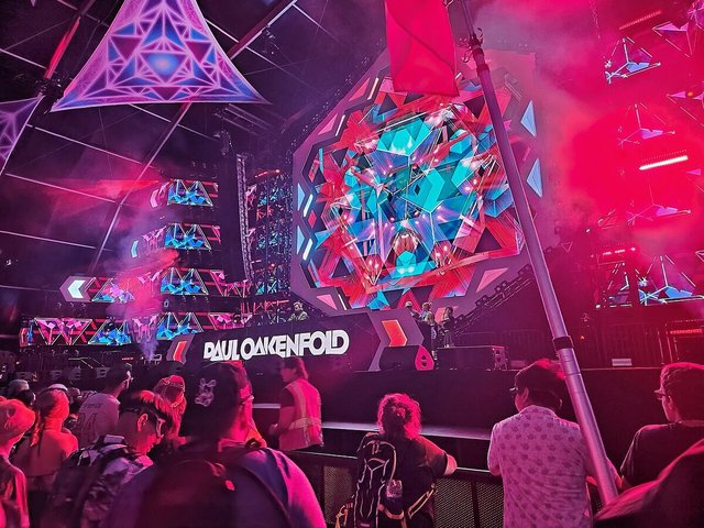 Paul Oakenfold name on a festival stage with colorful geometric video screens, seen over a crowd from behind