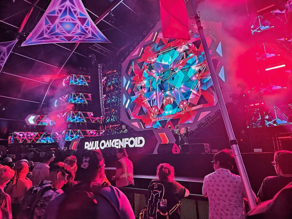 Paul Oakenfold name on a festival stage with colorful geometric video screens, seen over a crowd from behind