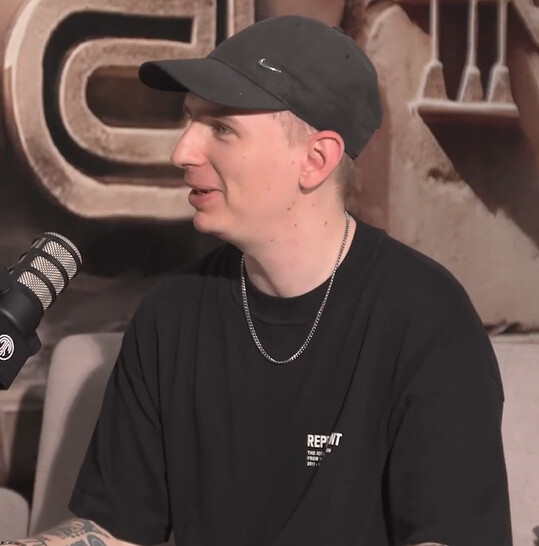 Klangkuenstler in a black cap and black T-shirt, seated beside a podcast microphone