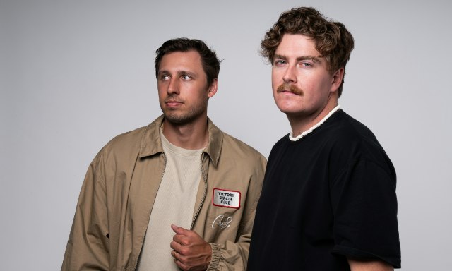 Two men posed against a plain gray backdrop, one in a tan work jacket and one in a black shirt