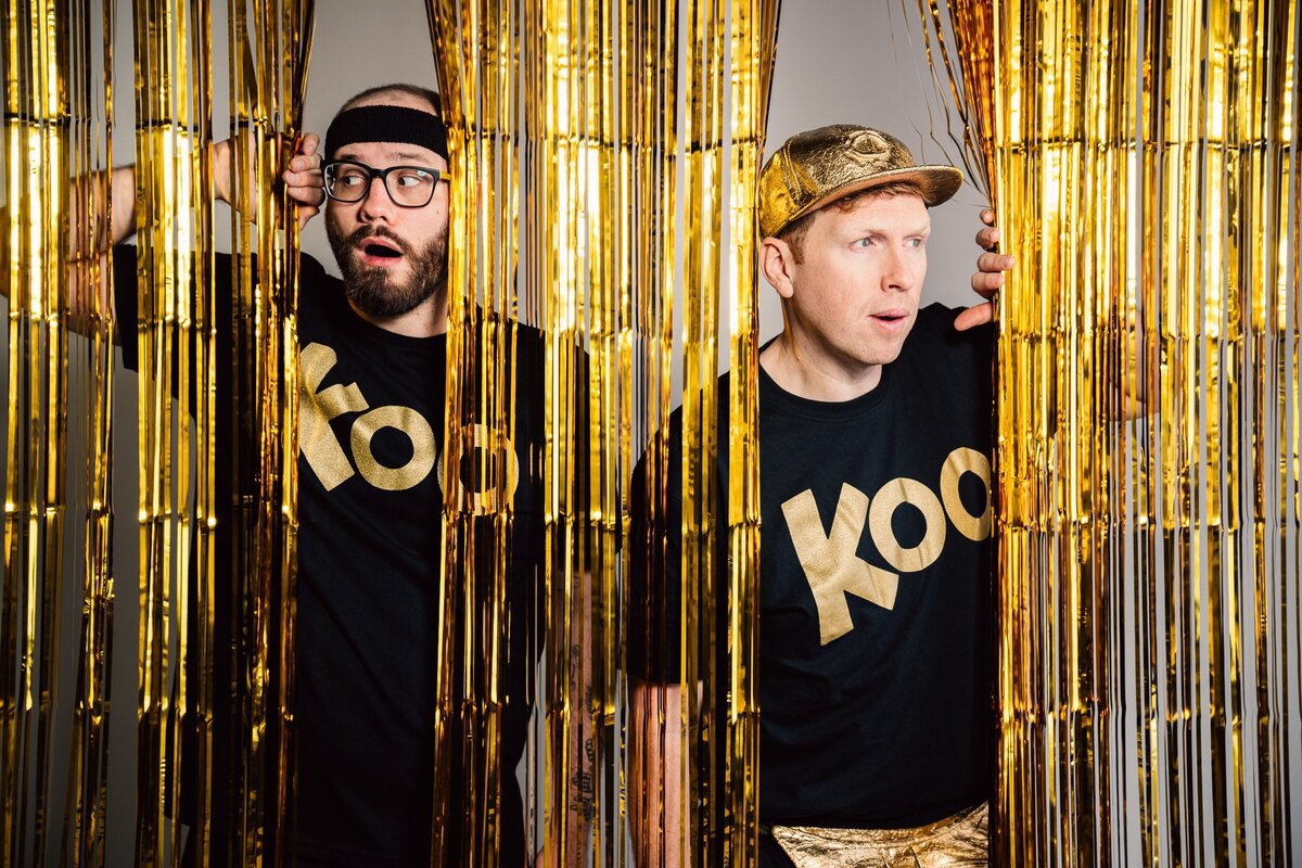 Two men in black Koo T-shirts, one in a headband and glasses, one in a gold cap, peeking through gold foil fringe curtains