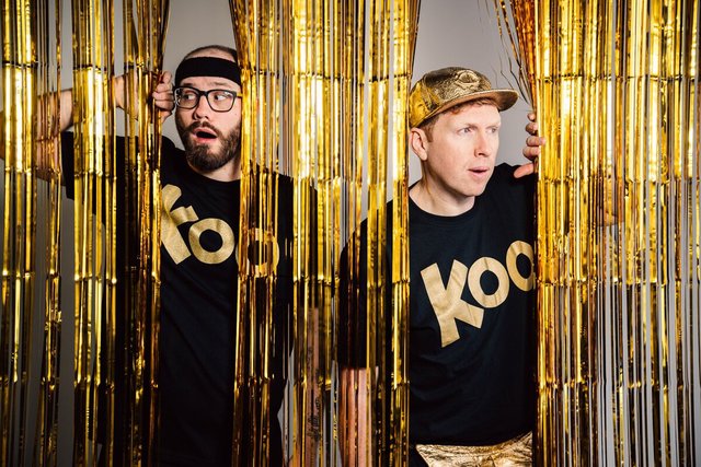 Two men in black Koo T-shirts, one in a headband and glasses, one in a gold cap, peeking through gold foil fringe curtains