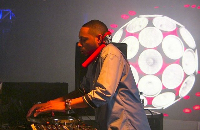 DJ in headphones working a mixer in front of a video screen showing a ball of speaker cones