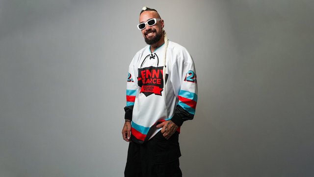 Lenny Pearce smiling in white sunglasses and a white hockey-style jersey printed with Lenny Pearce Toddler Techno, a long blond braid over one shoulder, against a gray backdrop