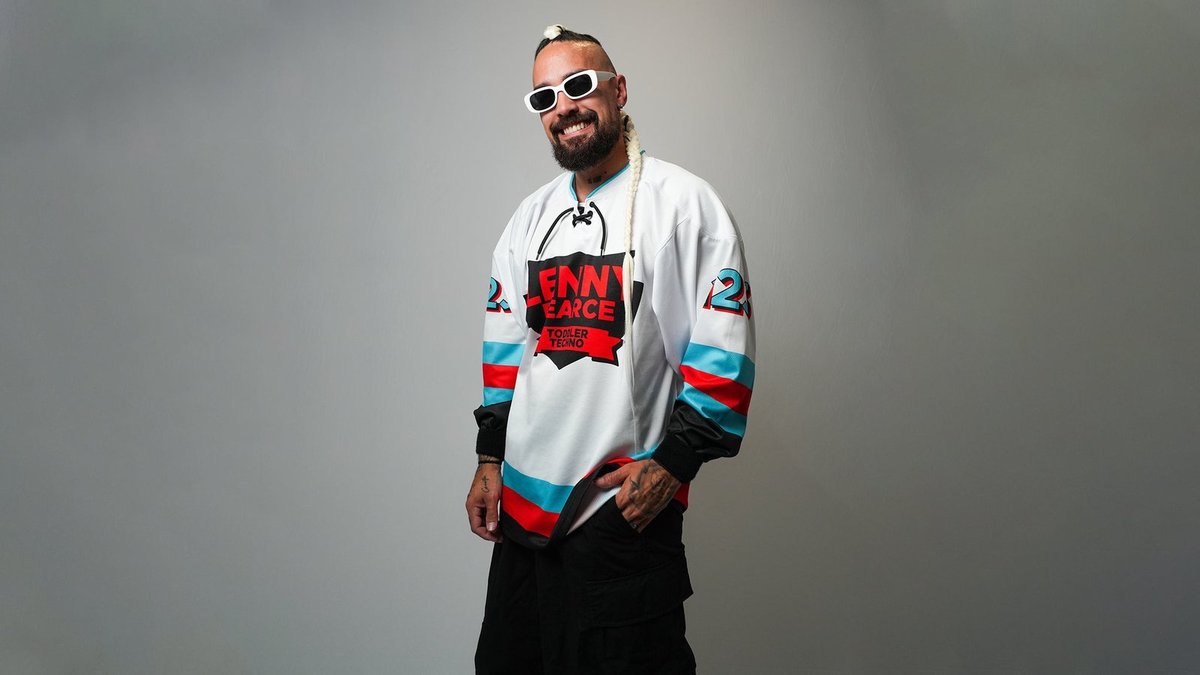 Lenny Pearce smiling in white sunglasses and a white hockey-style jersey printed with Lenny Pearce Toddler Techno, a long blond braid over one shoulder, against a gray backdrop