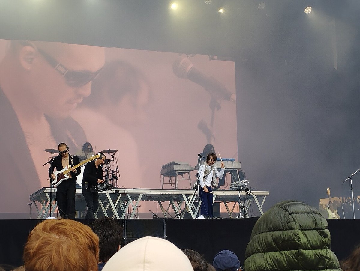 Musicians on an outdoor festival stage in front of a large pink video screen, with fog and stage lights above and the backs of heads in the crowd