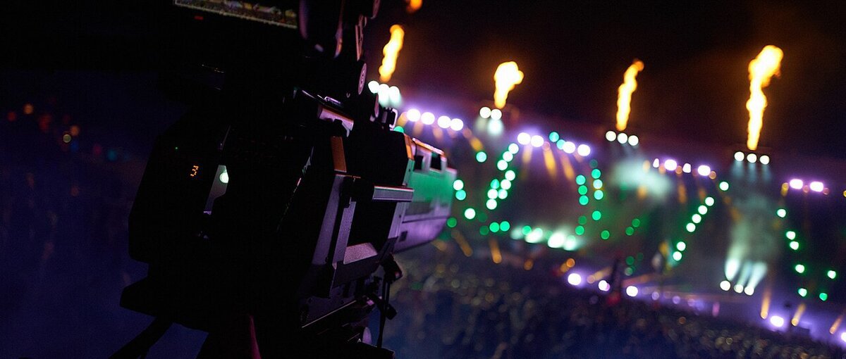 Festival main stage with flame jets and green and purple light rigs seen past a camera operator