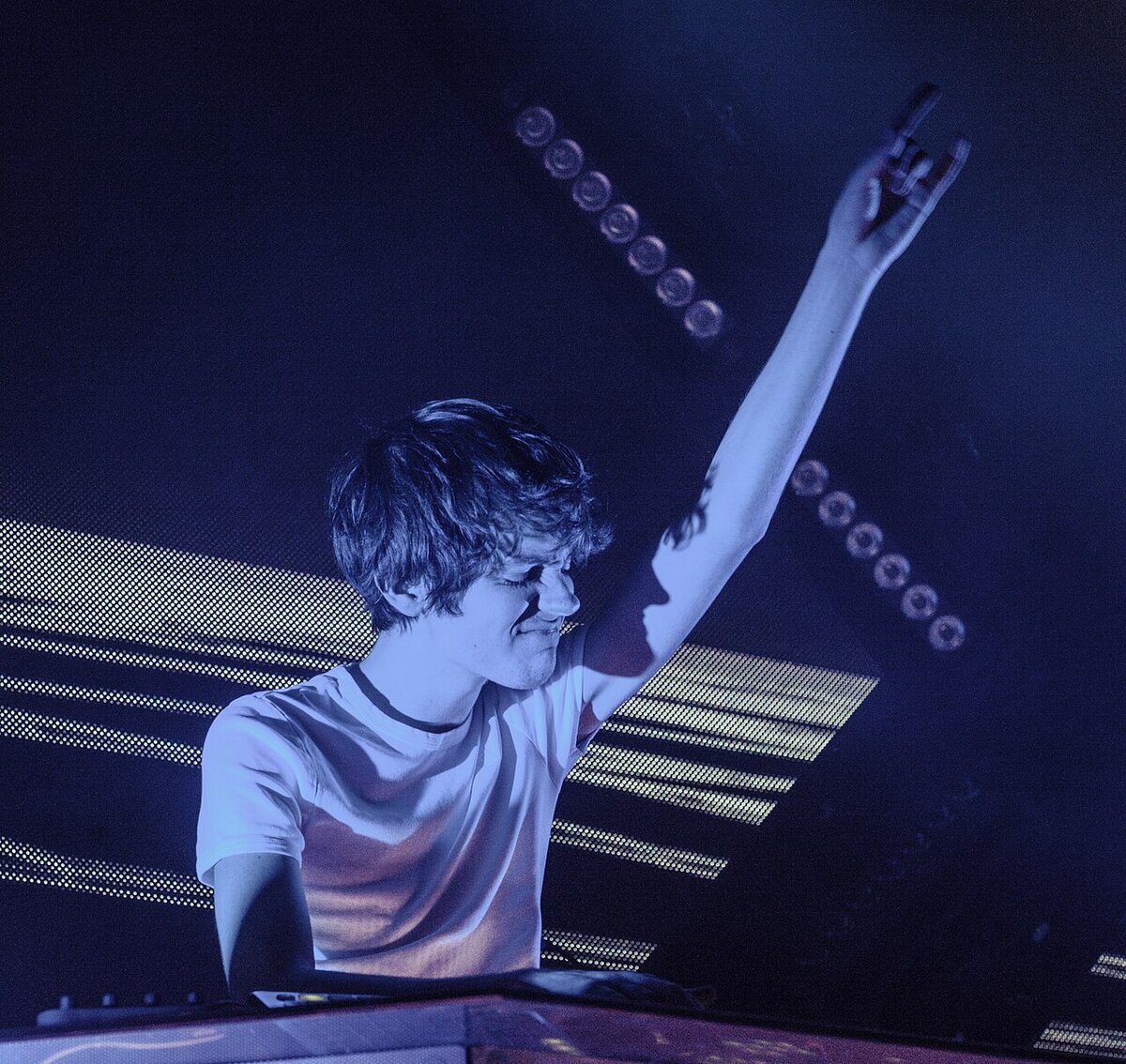 Madeon at the decks under blue stage lights, one arm raised