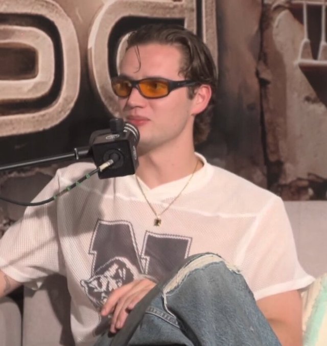 Mau P seated at a podcast microphone wearing orange-tinted sunglasses and a white mesh jersey