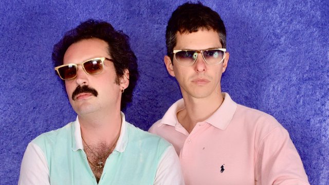 The two members of Mind Enterprises in tinted aviator sunglasses and pastel polo shirts against a textured purple backdrop